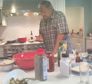 image of an old man looking at an ingredient he is holding while standing beside the kitchen table filled with seasonings